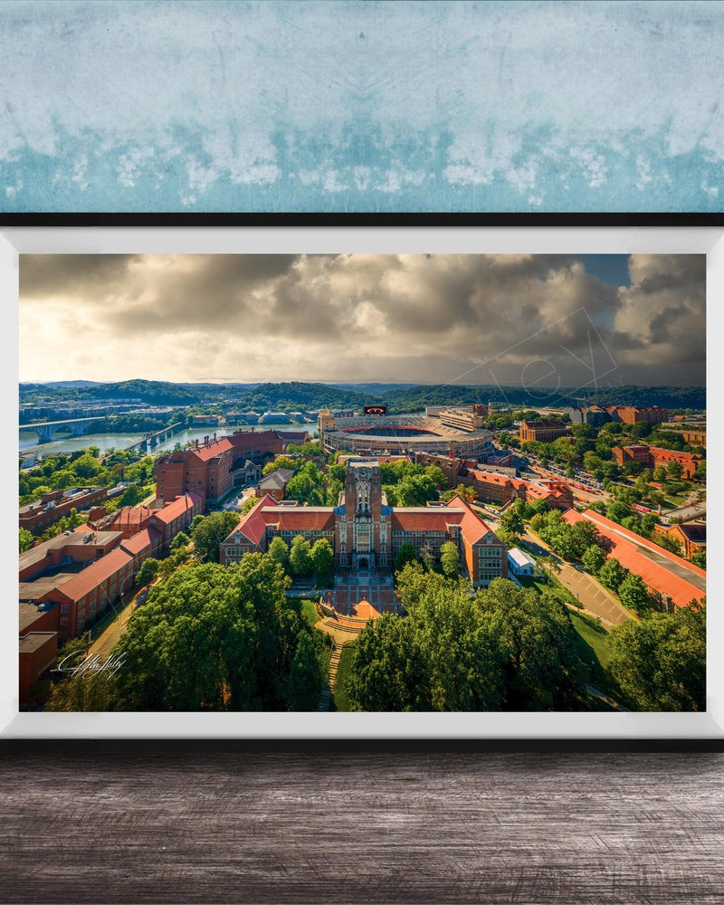 The image is a framed aerial view of a city with a mix of buildings, trees, and a river. The sky is cloudy, creating a dramatic backdrop for the cityscape.