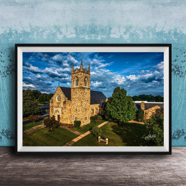 A stone church with a tall tower, surrounded by trees and a grassy area, set against a blue sky with clouds.