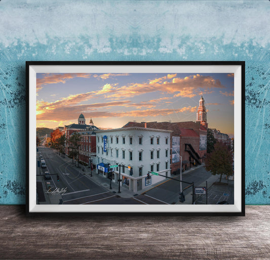 The image is a framed photograph of a city street at sunset, featuring a mix of architectural styles and a church with a tall tower.