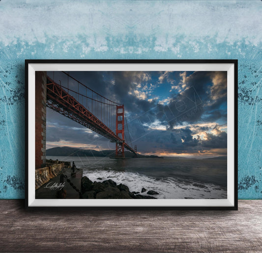 A framed photograph of the Golden Gate Bridge in San Francisco, California, with dramatic clouds and a sunset sky in the background.