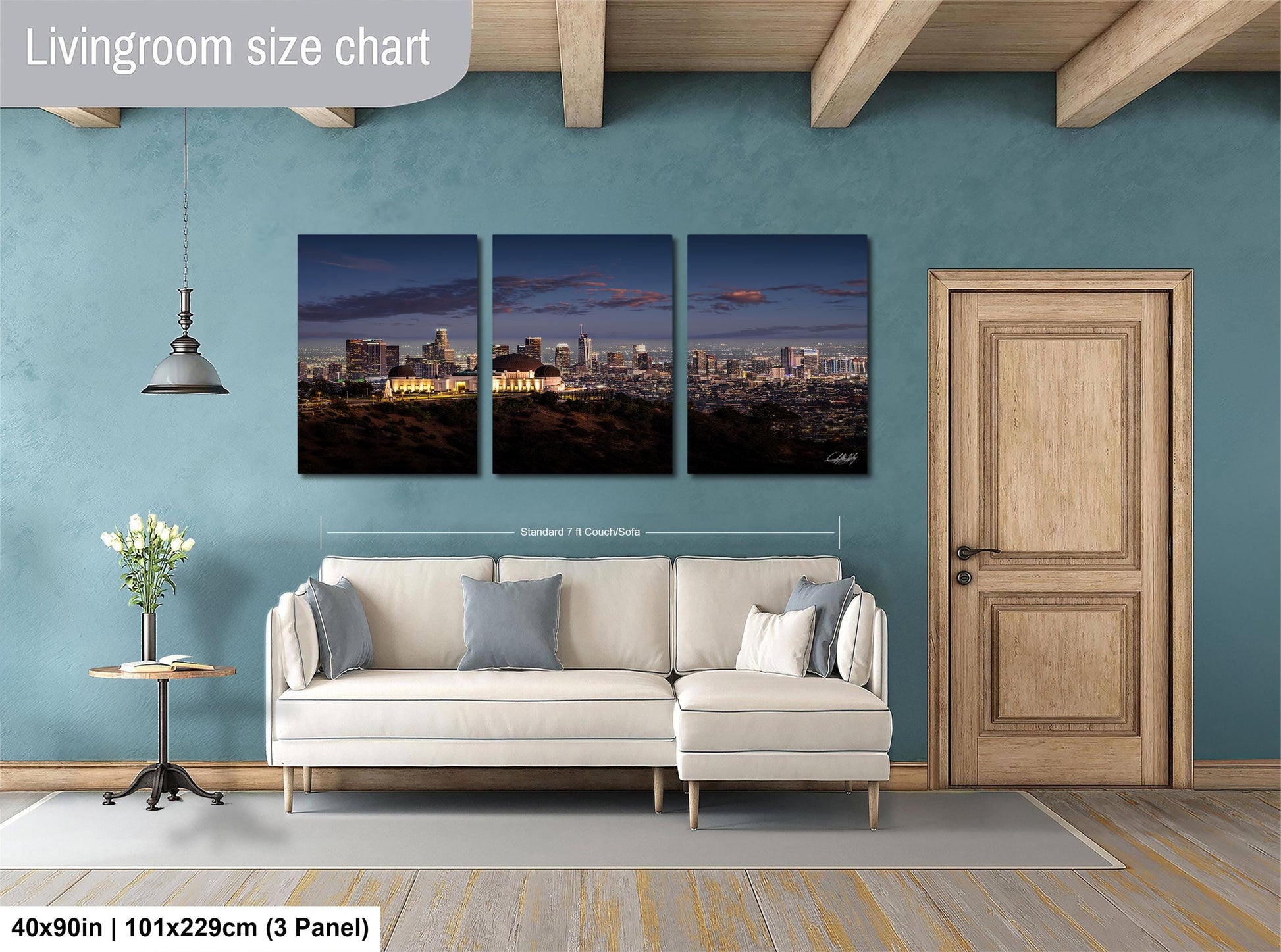 The image is a living room with a large wall art piece depicting a city skyline at night. The art piece is divided into three panels, each showing a different part of the cityscape. The living room has a white sofa and a wooden door, and the floor is covered with a gray rug.