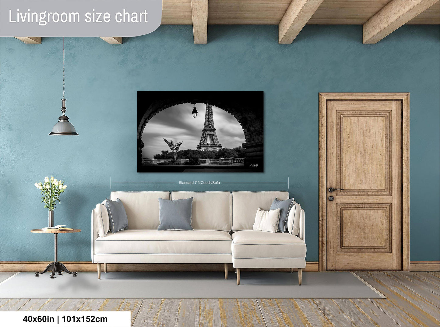 a living room with a large black and white photograph of the Eiffel Tower hanging on the wall. The living room has a white sofa, a wooden door, and a wooden table with a vase of flowers. The photograph is approximately 40 inches wide and 115 centimeters tall.