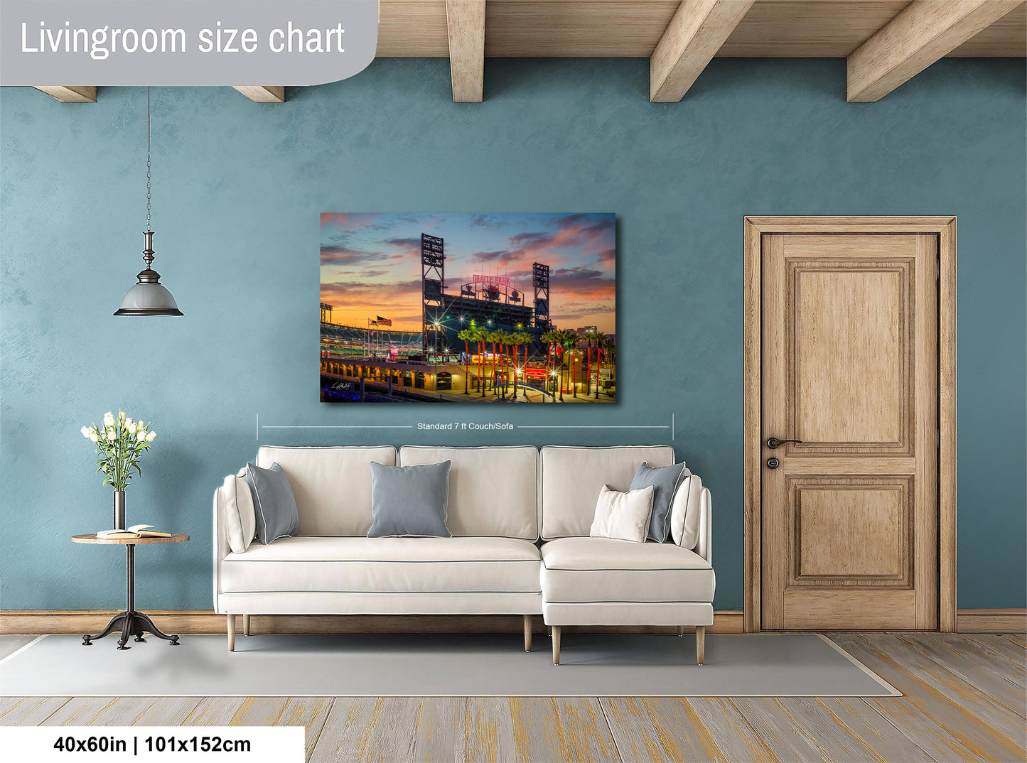 a living room with a large wall painting depicting a baseball stadium at sunset. The painting is hanging above a white sofa, which is positioned in front of a wooden door. The room has a wooden floor and a small table with a vase of flowers. The wall painting is approximately 40 inches wide and 115 centimeters tall.