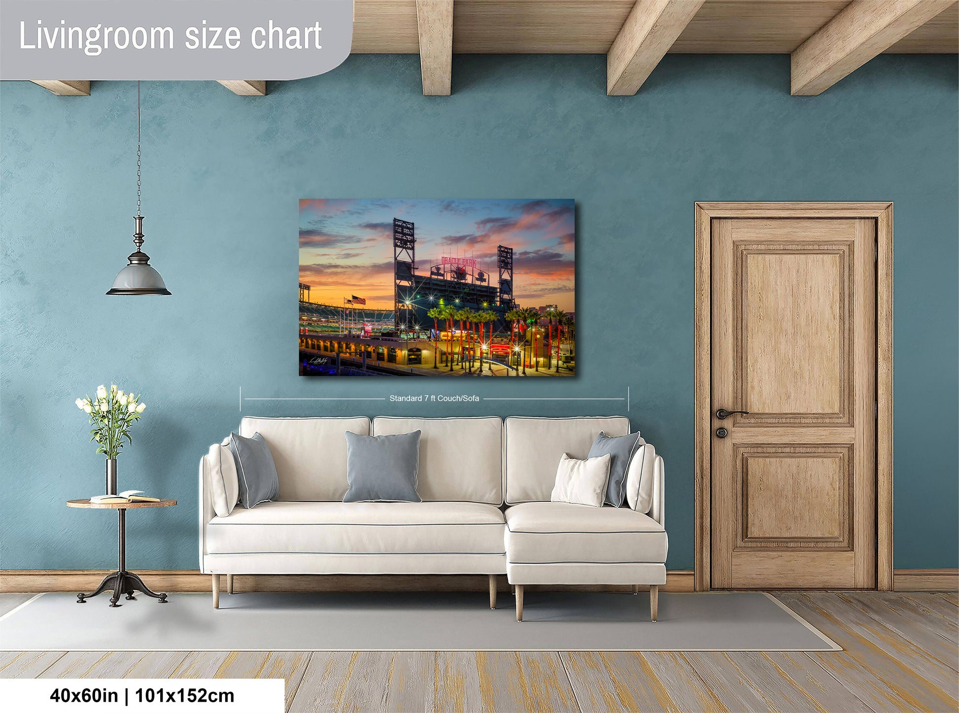 a living room with a large wall painting depicting a baseball stadium at sunset. The painting is hanging above a white sofa, which is positioned in front of a wooden door. The room has a wooden floor and a small table with a vase of flowers. The wall painting is approximately 40 inches wide and 115 centimeters tall.