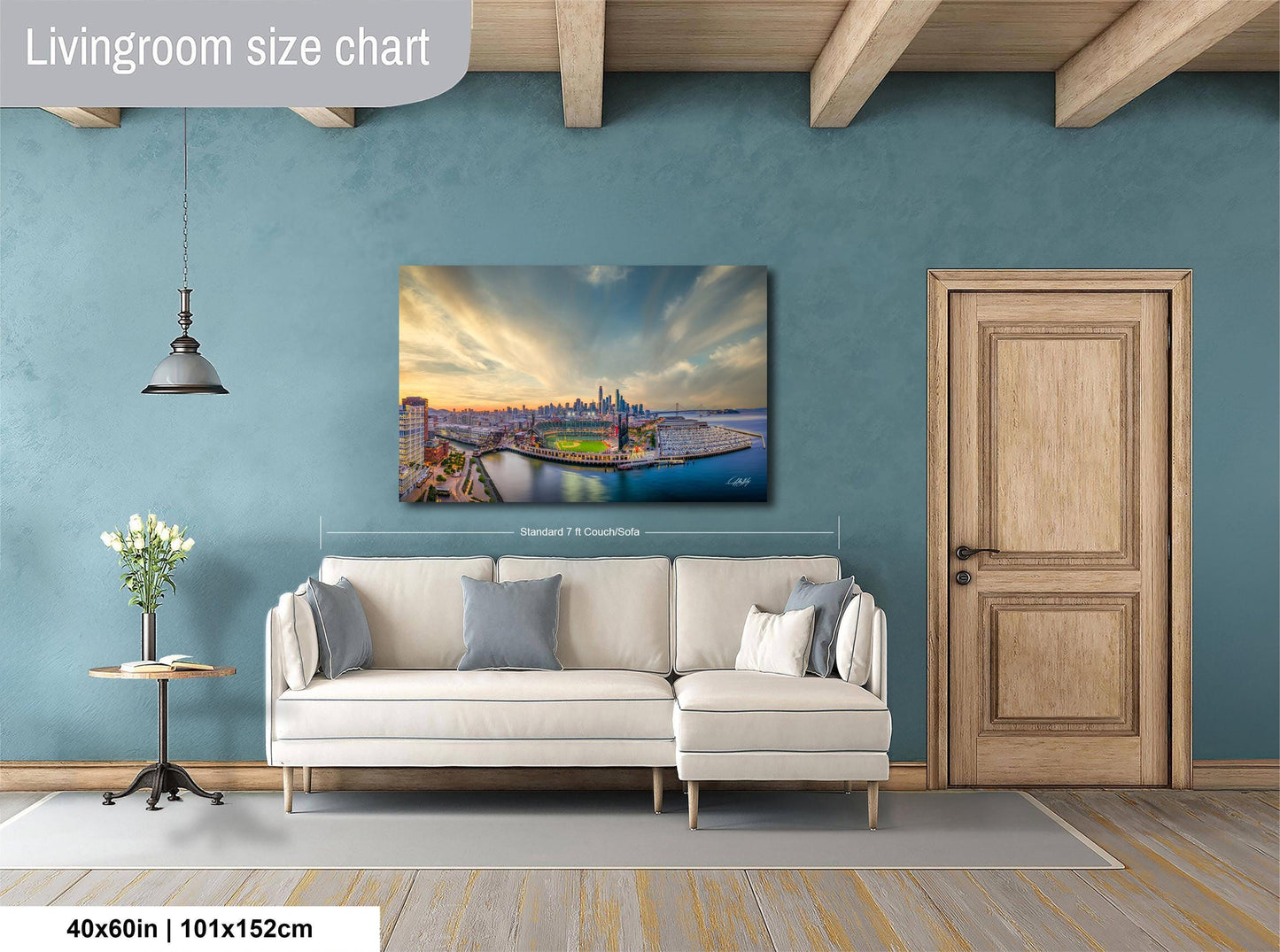 a living room with a large painting of a cityscape hanging on the wall. The painting is framed and measures 40 inches by 101.5 centimeters. The room has a white sofa, a wooden door, and a small table with a vase of flowers.