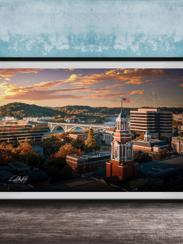 The image is a framed photograph depicting a cityscape with a bridge, buildings, and a clock tower, all set against a colorful sunset sky.