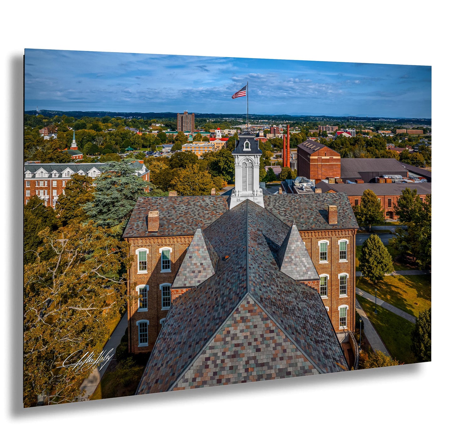 a large brick building with a steep roof and a flag flying atop it, surrounded by trees and other buildings in a cityscape.