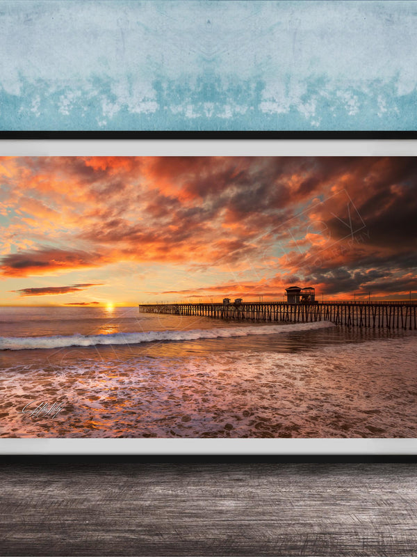 A framed photograph depicting a serene beach scene at sunset, with a wooden pier extending into the water and the sky filled with vibrant orange and pink hues.