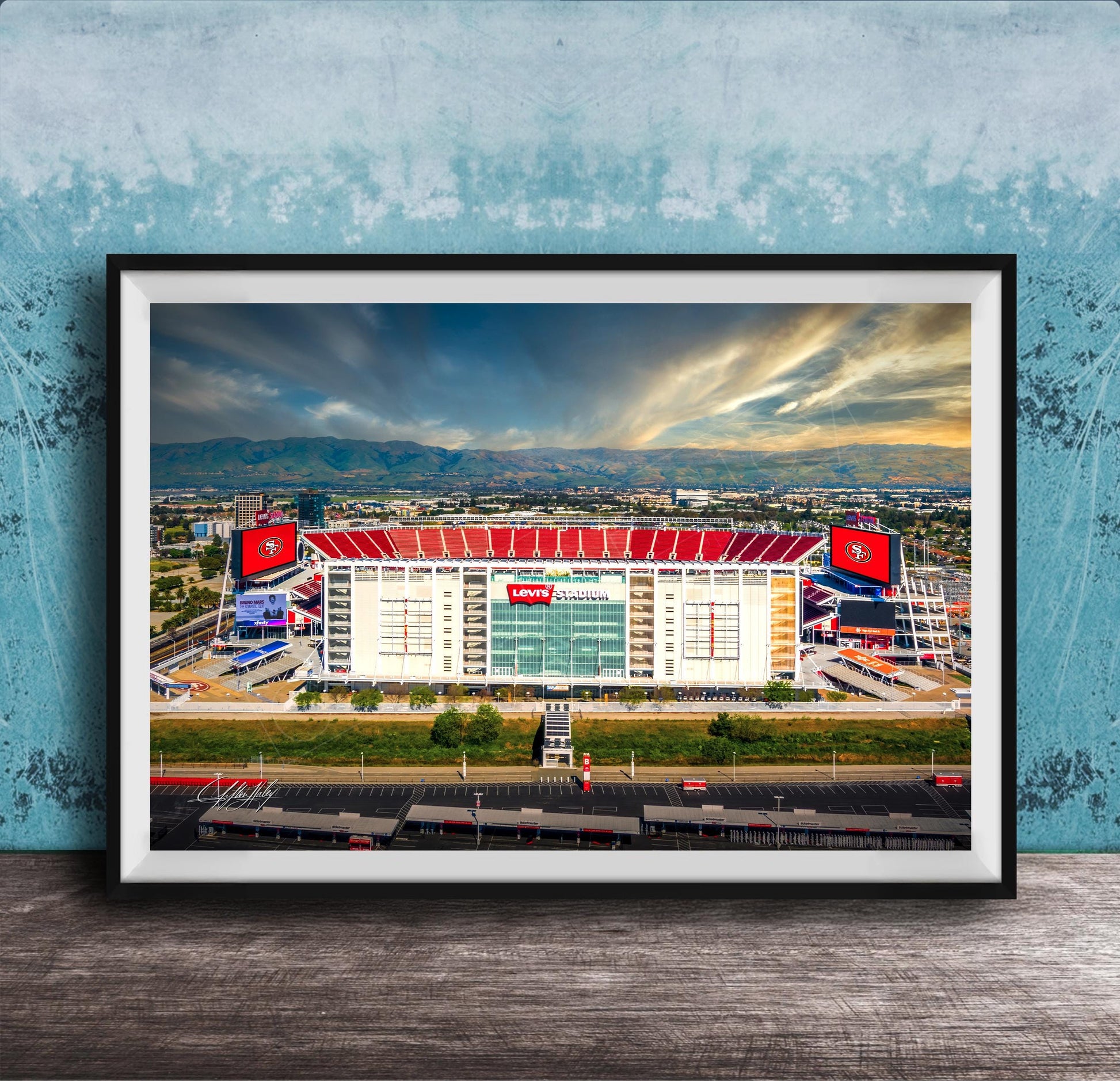 A framed photograph of a large sports stadium with a red roof, surrounded by mountains and a cloudy sky.