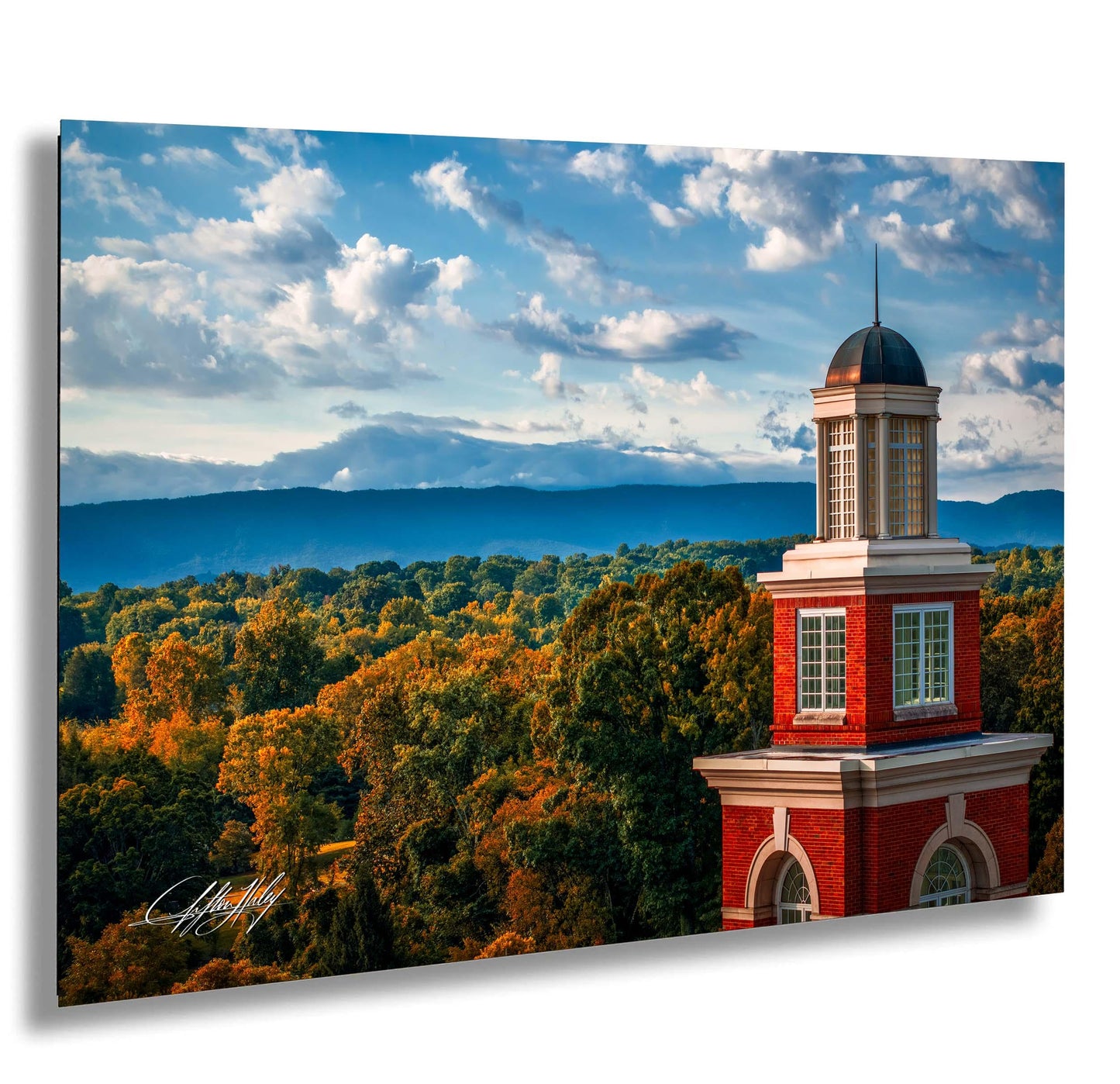 a red brick building with a dome and a tower, surrounded by a lush green forest with trees displaying autumn colors. The sky above is blue with white clouds.