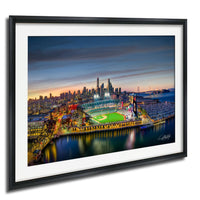A framed aerial view of a city skyline at dusk, with a baseball field and stadium illuminated in the foreground.