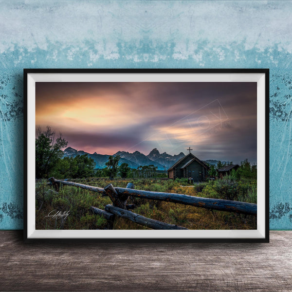 A framed photograph depicting a serene rural landscape with a wooden fence, a church, and mountains in the distance, set against a colorful sunset sky.