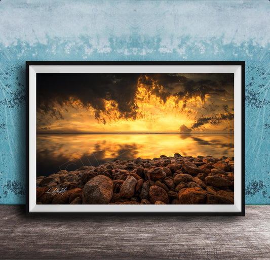 The image is a framed photograph depicting a rocky shoreline at sunset, with the sky filled with vibrant orange and yellow hues reflecting on the calm water surface.