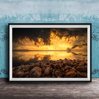 The image is a framed photograph depicting a rocky shoreline at sunset, with the sky filled with vibrant orange and yellow hues reflecting on the calm water surface.