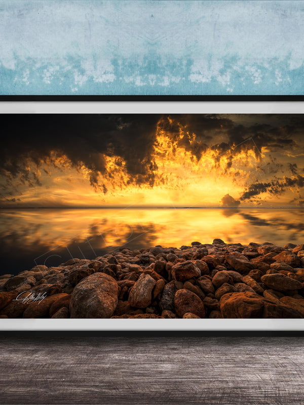 The image is a framed photograph depicting a rocky shoreline at sunset, with the sky filled with vibrant orange and yellow hues reflecting on the calm water surface.