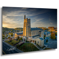 A large stone church with a tall tower, surrounded by a grassy area and a parking lot, set against a backdrop of a cloudy sky and a sunset.