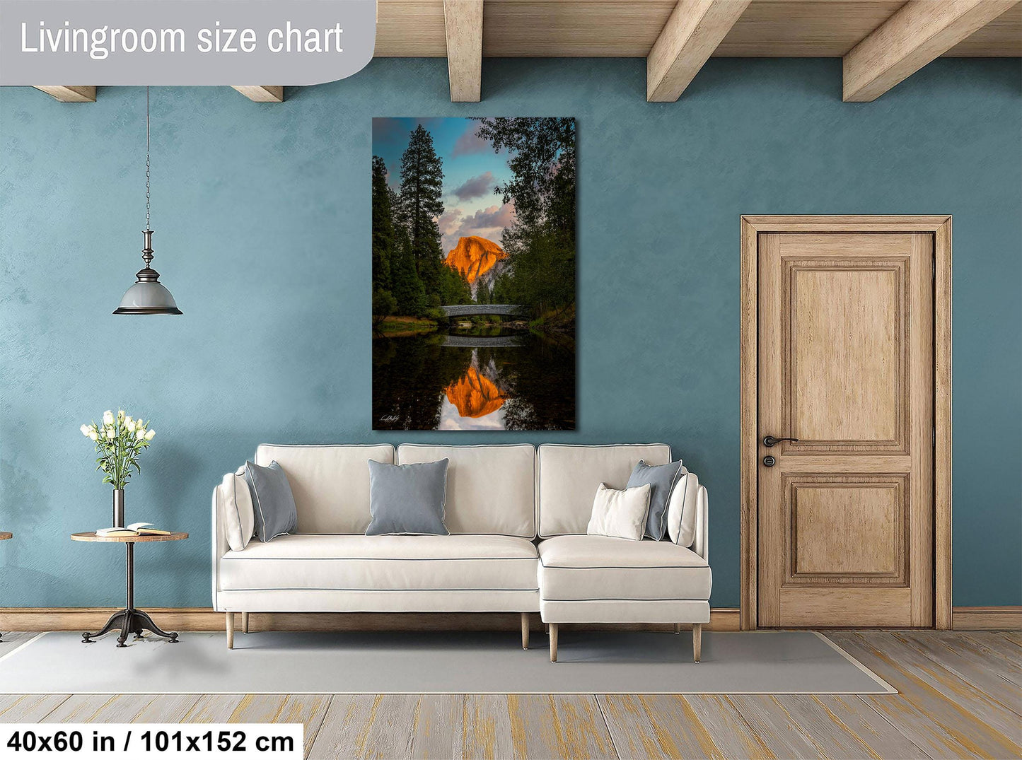 a living room with a large painting of a sunset over a lake hanging on the wall. The painting is framed in a white frame and is approximately 40 inches wide and 51 inches tall. The room has a white sofa, a wooden door, and a small table with a vase of flowers.