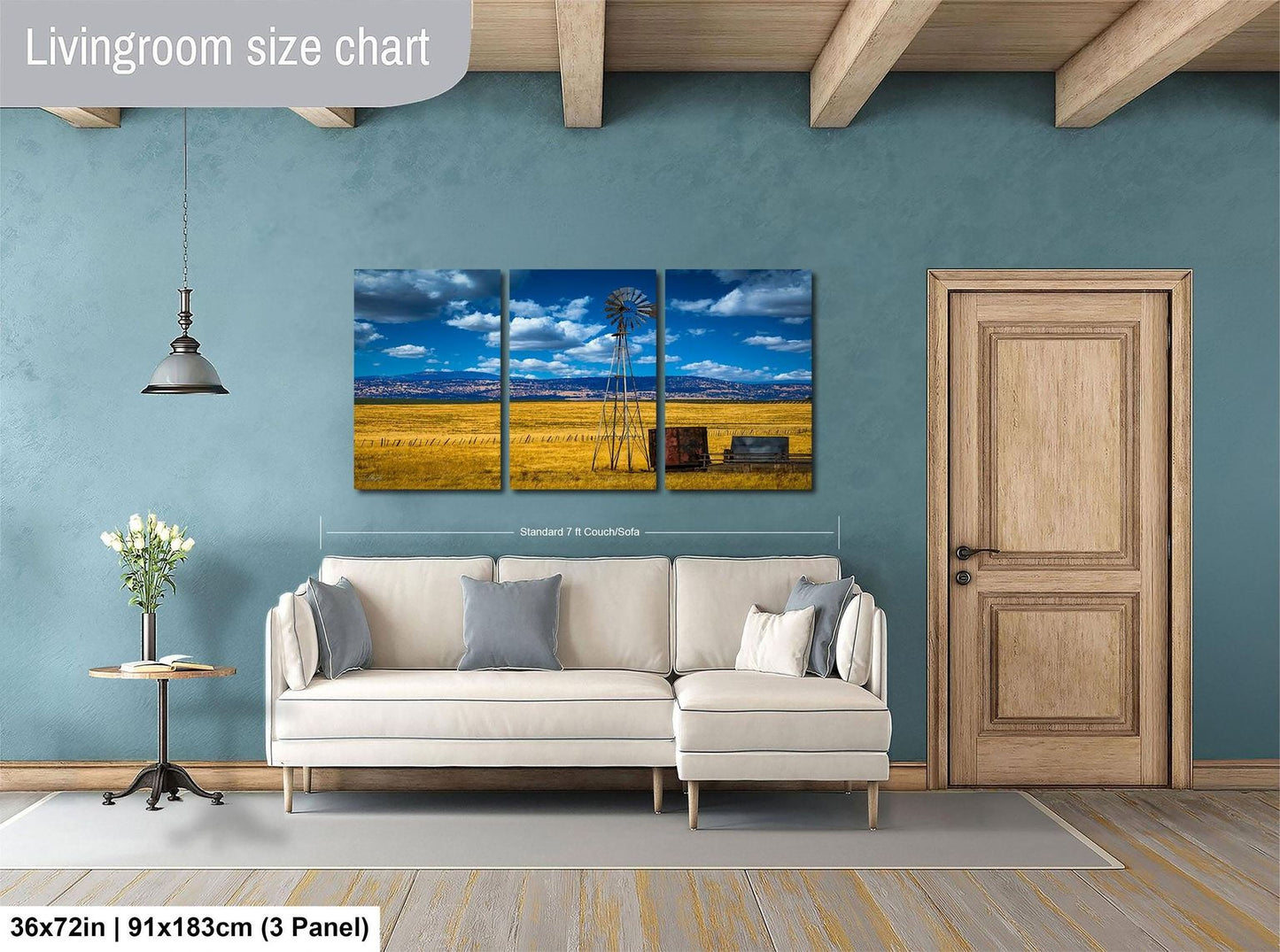 The image is a living room with a large, three-panel landscape painting hanging on the wall. The painting depicts a rural scene with a windmill and a field of crops. The room has a white sofa and a wooden door, and there is a vase of flowers on a table. The painting is approximately 36 inches wide and 91 inches tall, and it is mounted on a wooden beam.