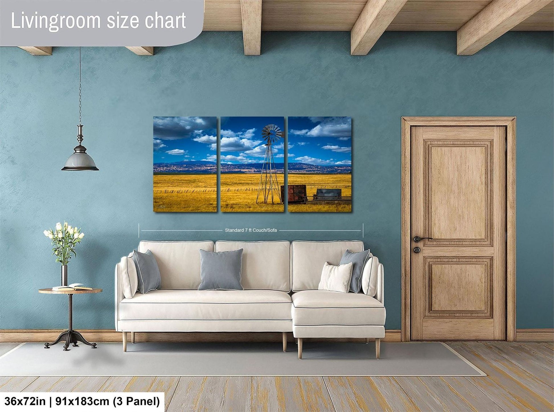 The image is a living room with a large, three-panel landscape painting hanging on the wall. The painting depicts a rural scene with a windmill and a field of crops. The room has a white sofa and a wooden door, and there is a vase of flowers on a table. The painting is approximately 36 inches wide and 91 inches tall, and it is mounted on a wooden beam.