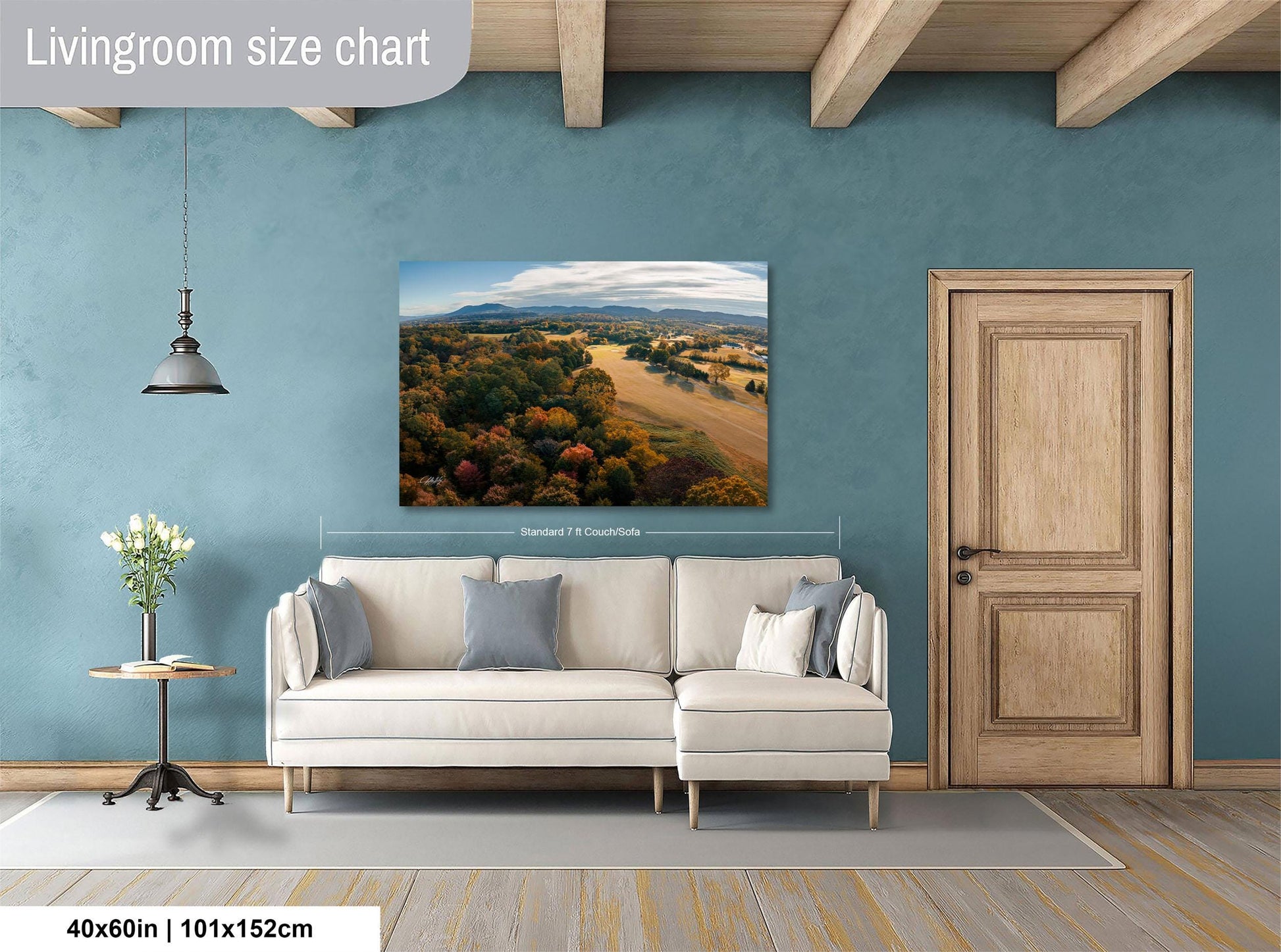 a living room with a large landscape painting hanging on the wall above a white sofa. The painting depicts a scenic countryside view with rolling hills, trees, and a blue sky. The room has a wooden door, a small table with a vase of flowers, and a wooden beam ceiling. The painting is approximately 40 inches wide and 115 centimeters tall.
