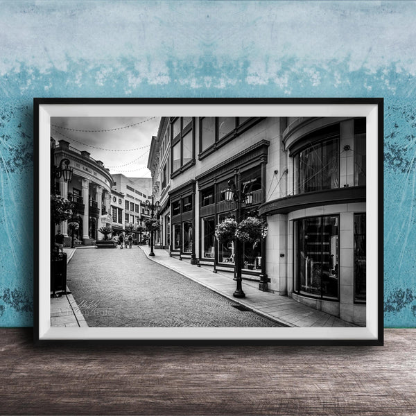 A black and white photograph of a city street lined with buildings and shops, with a cobblestone road and street lamps.