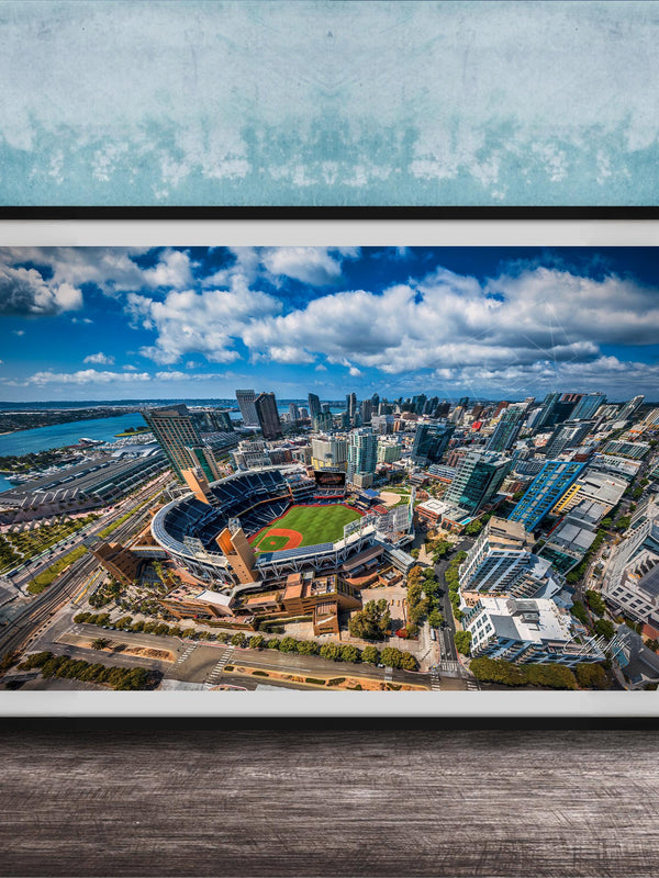 The image is an aerial view of a city with a baseball stadium in the center, surrounded by tall buildings and a body of water.