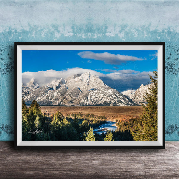 A framed photograph of a mountainous landscape with snow-capped peaks, a river, and a forested area in the foreground.