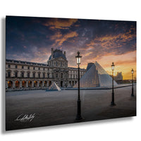 the Louvre Museum in Paris, France, with its iconic glass pyramid and surrounding architecture illuminated against a vibrant sunset sky.