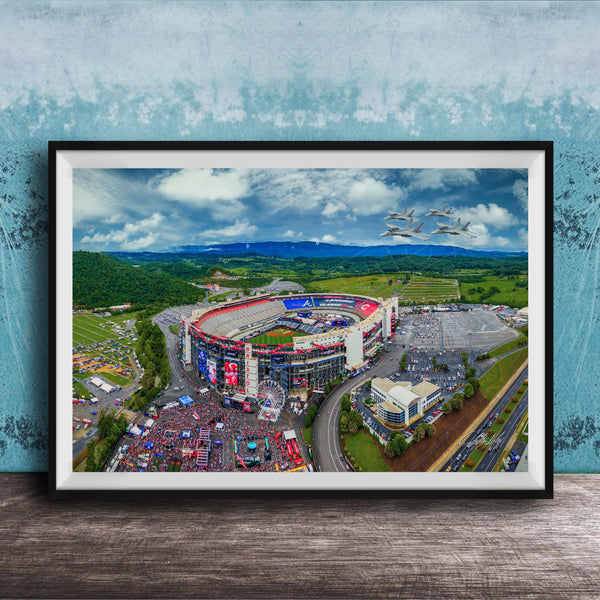 The image is an aerial view of a large stadium surrounded by a green landscape, with a few buildings and vehicles visible.