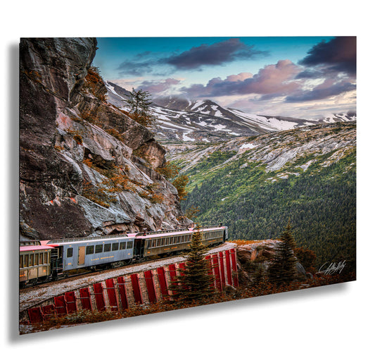 A train travels along a winding track that runs parallel to a steep, rocky cliff face, with snow-capped mountains visible in the background under a partly cloudy sky.