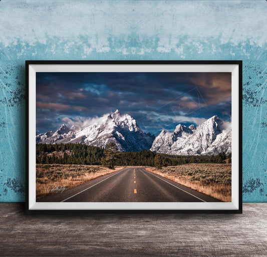 A framed photograph of a winding road leading towards majestic snow-capped mountains against a dramatic cloudy sky.