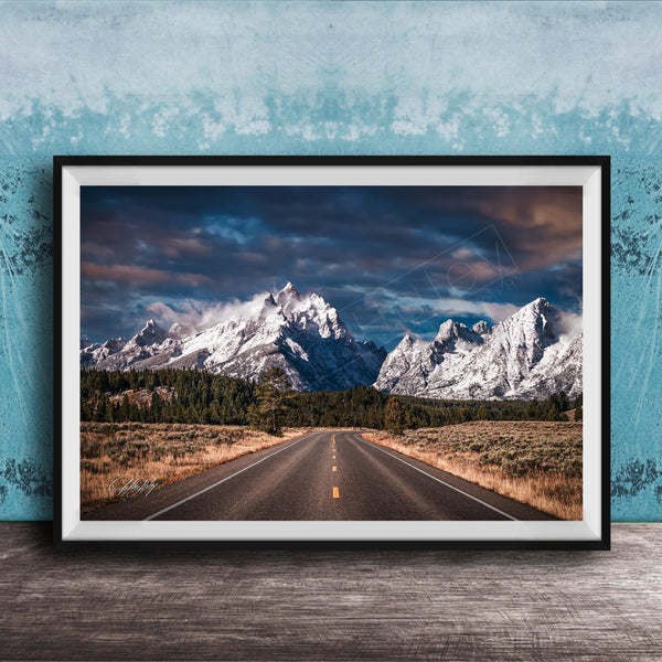 A framed photograph of a winding road leading towards majestic snow-capped mountains against a dramatic cloudy sky.