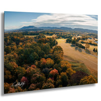 The image is a scenic aerial view of a countryside landscape with a mix of trees in autumn colors, rolling hills, and a distant mountain range under a blue sky with clouds.