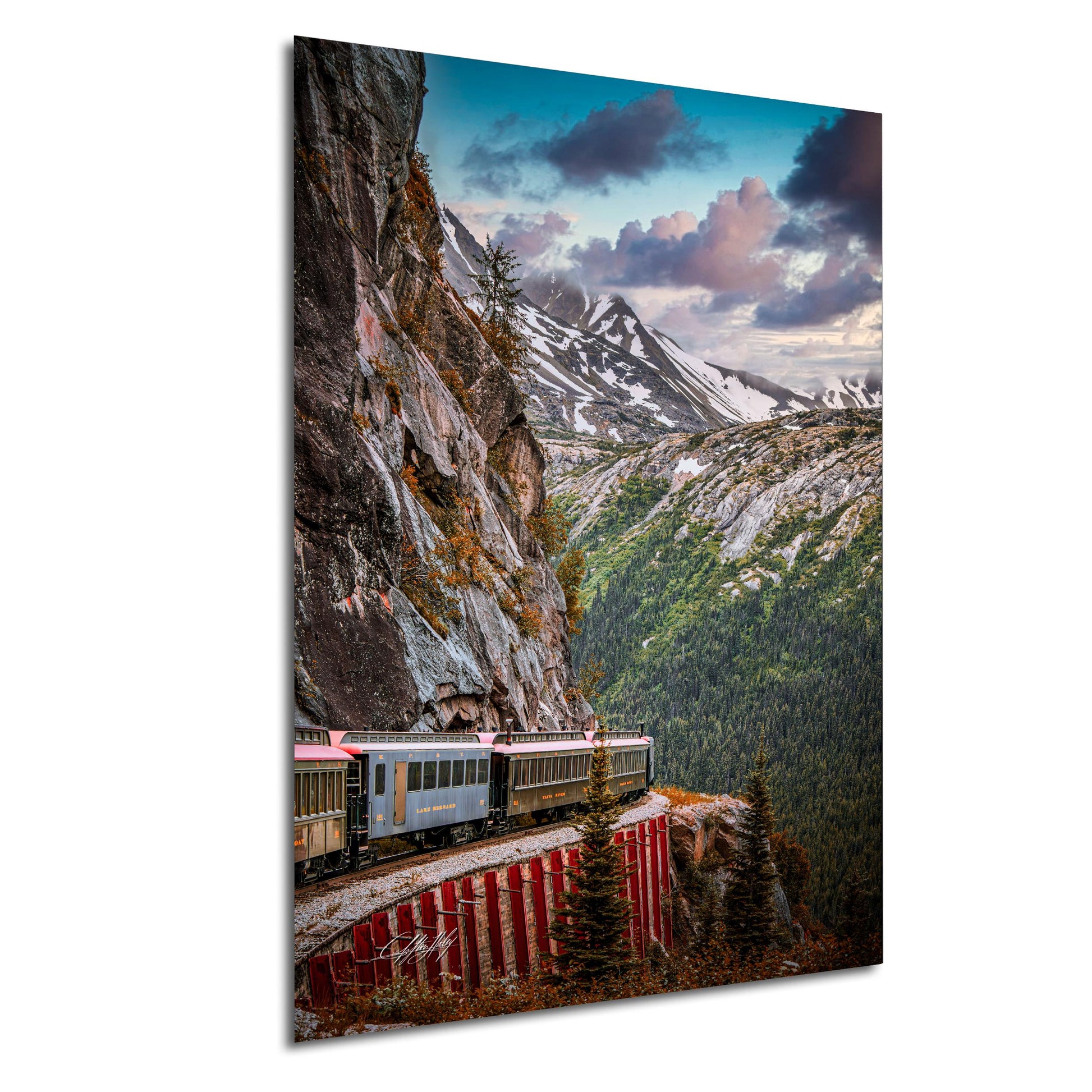 A train travels along a track that runs along the side of a steep, rocky cliff, with snow-capped mountains visible in the background under a cloudy sky.