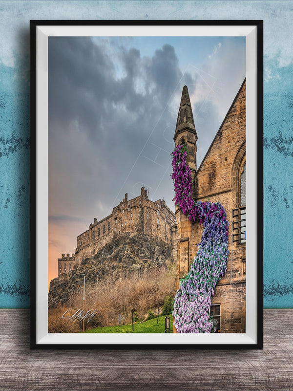 A framed photograph of a castle with a tower, set against a cloudy sky, with a colorful flower arrangement hanging from the tower.