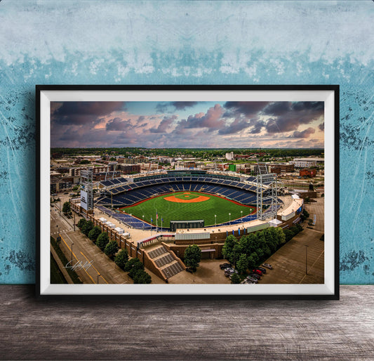 An aerial view of a baseball stadium with a lush green field, surrounded by buildings and a cloudy sky.
