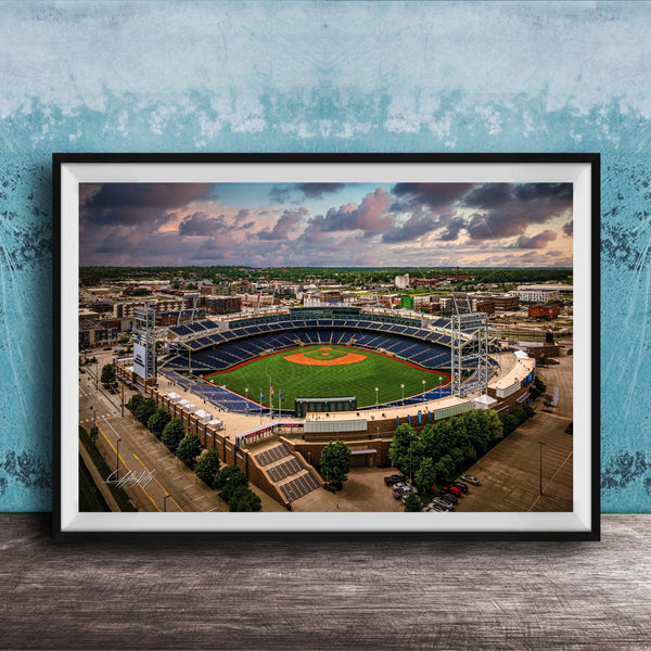 An aerial view of a baseball stadium with a lush green field, surrounded by buildings and a cloudy sky.