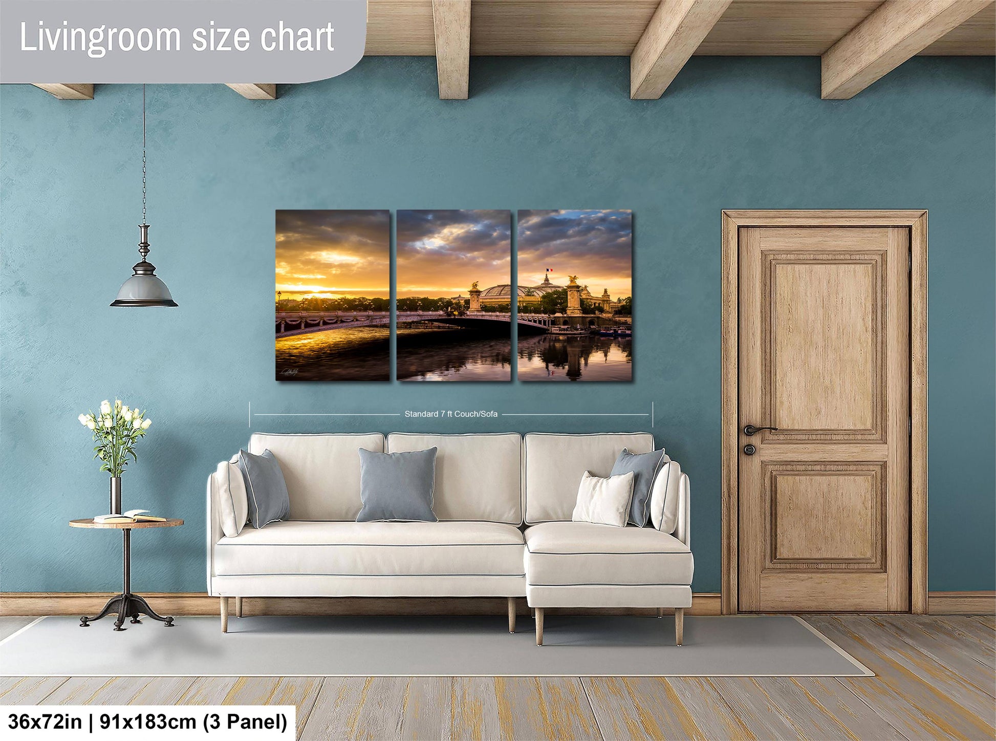 The image is a living room with a large, three-piece wall art depicting a scenic view of a bridge and a sunset. The wall art is mounted above a white sofa, and there is a wooden door and a small table with a vase of flowers nearby. The living room appears to be well-lit and spacious, with a wooden floor and a blue wall.
