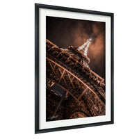 The image is a framed photograph of the Eiffel Tower, a famous landmark in Paris, France. The photograph is taken from a low angle, emphasizing the tower's height and intricate design. The sky in the background is dark, creating a dramatic contrast with the illuminated structure. The image is framed within a black border, highlighting the architectural details of the Eiffel Tower.